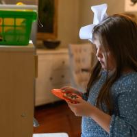 Henley stands in a play kitchen looking down at a cookie on an orange plate that she is holding.