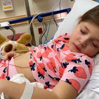 Henley lays in a hospital bed with different wires attached to her. She is wearing pink pajamas with strawberry designs on them.