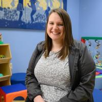 Sarah McAlister, a white adult woman with brown hair, smiles as she sits in the Child Life room at Kentucky Children’s Hospital, surrounded by toys and art. She is wearing a light grey top with a dark grey jacket.