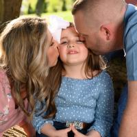 Henley smiles as she sits in between her mom, Lindsay, a middle-aged white woman with blonde hair, wearing a pink top, and her father, Jarred, a middle-aged white man with short brown hair, wearing a blue shirt. They kiss either side of her cheeks.