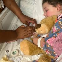 Henley and Donnie snuggle in a hospital bed covered by blankets as a nurse checks on Henley’s IV.