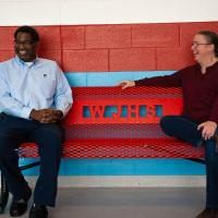 Ivan and Marci sit on a bench with WJHS on it. They are smiling and laughing.