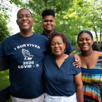 The Johnnson family stands in front of a black plank fence. Jerome has his arm around Jeanine’s shoulders. He is wearing a shirt that says “I survived 2020 COVID-19.”