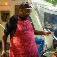 A candid photo of Joshua dipping his brush in a container with his sauce in it that a member of the Big Daddy cooking team is holding. The member of the cooking team is an older Black man with a gray beard. He is wearing a baseball hat that reads “Big Daddy’s BBQ” and a black short-sleeve t-shirt.