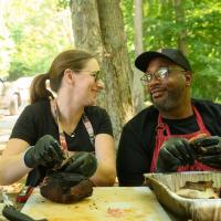 A photo of Joshua and Jill smiling at each other as they’re rubbing sauce on meat.