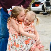 Wearing matching dresses, Julie embraces her younger sister from behind and they smile together an outside setting.