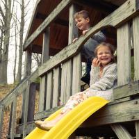 Julie and her younger brother sit in a treehouse smiling at the camera and people on the playground.