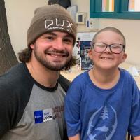 Kash Daniels, a football player, poses for a picture next to a little girl, Ellie. Ellie has buzzed hair and glasses. Kash is wearing a brown toboggan. Both are smiling.