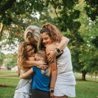 Kelley hugs her girls tightly. They are standing in a park. All are smiling.