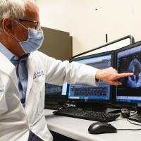 Dr. Booth points to the screen displaying the echocardiogram. He has a face mask over his nose and mouth.