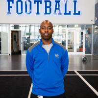 Gabe Amponsah, a bald black man, stands with his hands behind his back. He is wearing a blue and grey UK sweatshirt.