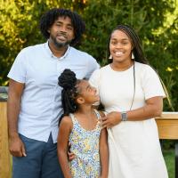 Kristen, her mother, and her father, LaMontrose, an African-American man with dark medium-length curly hair, smile and pose for the camera together. He is wearing a short-sleeve white button-up and a pair of blue shorts.