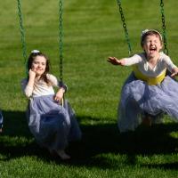 Leorah and her siblings play on their swingset, with Lenorah and her younger brother swinging while laying on their stomachs. Her brother is a young white boy, younger than his sisters, and is wearing a blue button-up and brown slacks. Her sister is a young white girl with brown hair, and is wearing a dress made of lace at the top and light blue tulle at the bottom.