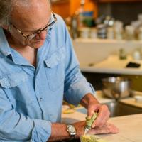 Lynn Sweet, an older man with gray hair and horn-rimmed glasses, does some detail work on a painting. He is wearing a blue button-up shirt.