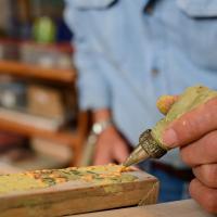 A close-up of Lynn’s hand, holding a piping bag with a metal tip on it. A thick orange substance that looks like paint is being squeezed out of the piping tip onto the edge of a canvas.