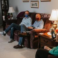 Mae sits on a single couch with her facemask, on a video call with her daughter-in-law, a young white woman with a big smile. Spread across the room are three middle-aged men sitting cross-legged with their facemasks on.