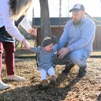 MaKenzie sits outside on a swing, flanked by her mother, Felicia, and her father, Timothy, a middle aged white man. He is wearing a light blue sweater, a grey and white baseball cap with blue jeans.