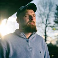 Timothy stands on his porch as the sun sets beneath the trees behind him. He is looking past the camera with a determined expression.