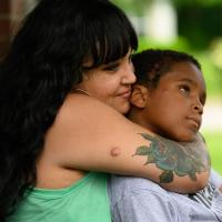 A candid photo of Malakai’s mother, Cacy Roberts, smiling as she wraps her arms around Malakai. She is a Black woman with long dark hair and bangs. She is wearing a light green top and gold hoop earrings.