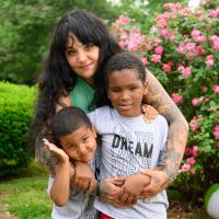 A photo of Cacy Roberts and her two boys posing together for the camera in front of a pink-flowering bush.