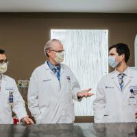 Marshall stands beside two directors of medical physics in his Radiation Oncology department: Dr. Damodar Pohkrel, middle-aged South Asian man with black hair, and Dr. Dennis Cheek, a middle-aged white man with grey hair. Both men are wearing their doctor’s coats and spectacles. All three are wearing masks.