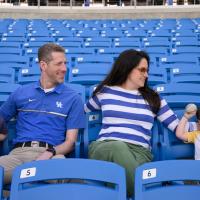 A candid photo of Matt, his wife Carmen, and their youngest son sitting in the stands of the stadium. Carmen is a white woman with long black hair. She is wearing a blue and white long-sleeve top with green pants, and a brown pair of tinted sunglasses. Their son is a young white boy with long brown hair. He is wearing a white and yellow short-sleeve shirt with Batman on it.