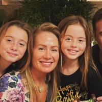 A family photo of Coach Mitchell with his wife and daughters. His daughter on the left is a young white girl with long blonde hair. She is wearing a black and white dress with a star pattern. His daughter who is second from right is a young white girl with long blonde hair, wearing a black and gold shirt. His wife (center) is a white woman with long blonde hair, wearing a floral pattern top.