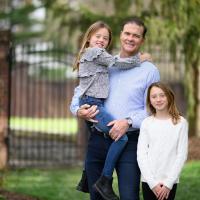 A photo of Coach Mitchell posing outside with his daughters, one of whom he is holding.