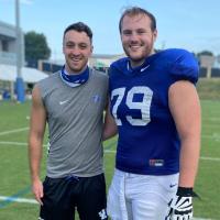 Luke and Max pose for a photo on the field. Both are wearing their practice gear and are smiling.