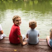 Max and his siblings sit on a dock. Max is turned to smile at the camera. His siblings are looking out at the lake.