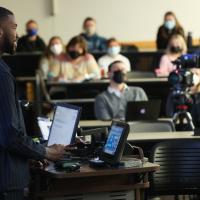 A candid photo of Michael giving a presentation while standing at a podium at the front of a classroom.