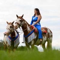 Miko sits on one horse, while the other horse stands next to them. The photo, taken in a field, is shot from a low angle, looking up at Miko. She is looking away from the camera.