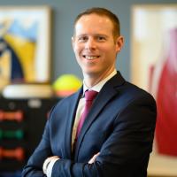 A photo of Dr. Austin Stone, a young-looking white man wearing a suit and smiling. He is standing in a physical therapy facility.