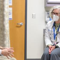 A photo of Dr. Sandra Beck and Ronnie Wu sitting in a doctor’s office while having a conversation. Dr. Beck is an older white woman with short white curly hair. She is wearing a white lab coat over a green shirt, several silver necklaces that have beads on them, and a pair of dark-rimmed glasses. Ronnie is a young Chinese man with short black hair. He is wearing a long-sleeve blue and white checkered button-up shirt with the sleeves rolled up and a pair of dark-rimmed glasses.