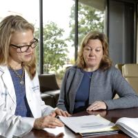 A candid photo of Dr. Kolesar and her colleague, Dr. Rachel Miller, sitting at a table together while looking at a piece of paper. Dr. Miller is a young white woman with long blonde hair. She is wearing a white lab coat over a blue top with a gold necklace, and a pair of dark-rimmed glasses.