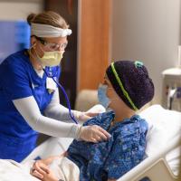 A photo of a nurse holding a stethoscope up to the chest of a patient laying in their hospital bed.