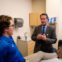 Peter props his knee up on an examination table while a Dr. Darren Johnson discusses treatment with him.