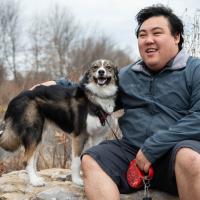 Roger sits outside on a large rock with his dog Pixel standing beside him on the rock.