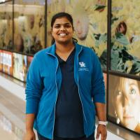 A photo of Dr. Rani Vasireddy smiling while standing in front of a wall decal with sunflowers on it. She is a South Asian woman with long black hair that is tied back in a ponytail. She is wearing a blue UK HealthCare-branded jacket over a pair of dark blue scrubs.