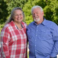 Ronnie and Becky pose together as they smile for the camera, with the treeline in the background.