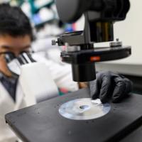 A close-up of Ronnie in a lab coat and set of gloves examining a slide underneath a microscope. The microscope is in focus, while he is slightly out of focus in the picture.