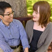 Ronnie and his girlfriend sit on a bench outside and smile at each other as they hold hands. She is a young white woman with light-brown hair in a mid-length cut, and she is wearing a grey sweater over a blue and white polka dot dress.