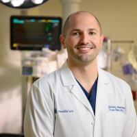 Dr. Zach Warriner, a white adult man with a shaved head, smiles while standing in front of medical equipment. He is wearing a white lab coat with a set of blue scrubs underneath.