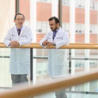 Dr. Zhonglin Hao, an older East Asian man with short greying hair, and Dr. Janeesh Sekkath Veedu, an older South Asian man with black hair, talk as they stand on a walkway in front of large glass windows. They are both wearing white lab coats with blue button-up shirts underneath. Dr. Hao is wearing glasses and a maroon tie, while Dr. Veedu is wearing a blue and white striped tie.