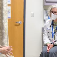 Ronnie sits and talks with Dr. Sandra Beck, an older white woman with a gray, curly, mid-length cut bob. She is wearing a white lab coat over a green sweater, a pair of grey slacks, and a stethoscope around her neck.