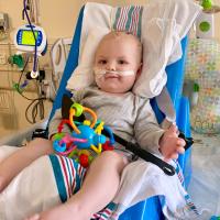 Rory is laying in his hospital bed with a colorful toy on his lap. He is wearing a grey striped outfit and has a tube in his nose.