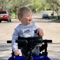 Rory stands smiling in an outside setting with the support of his gait trainer. An assistive medical device designed to train him in his walking pattern and assist with slow progressive weight bearing. He is wearing a grey lion sweatshirt with matching sweatpants.