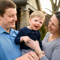 Rory smiles as he is held up in between his mother, Betsey—a young-looking, brunette white woman—and his father, Tyler—a young-looking brunette white man. Both parents are laughing as they face Rory. They are posing outside their home.