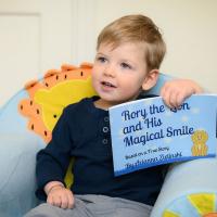 Rory sits in a light blue chair with a lion on it, and holds up a book: Rory the Lion and His Magical Smile.