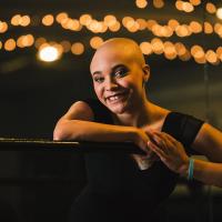 Savannah leans against a railing in her dance leotard and smiles. Her IV port is visible on her chest.